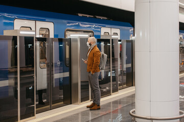 A man in a face mask to avoid the spread of coronavirus is holding a smartphone while waiting for a modern train at the subway platform. A bald guy in a surgical mask is keeping social distance.