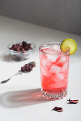 Refreshing hibiscus herbal tea made from magenta pink colored flowers served in drinking glass with ice cubes and lime slice on white wooden background with summer sunlight and shadows. Vertical