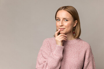 Curious sly woman millennial gentle smiling touching lips with finger. Cute blonde caucasian young female in casual pink knitted sweater and daily makeup isolated over grey studio wall background