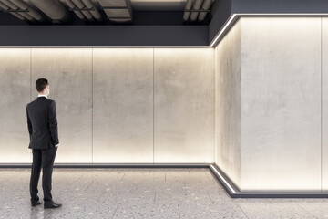 Businessman standing in modern exhibition space concrete interior with mockup place on wall. Mock...