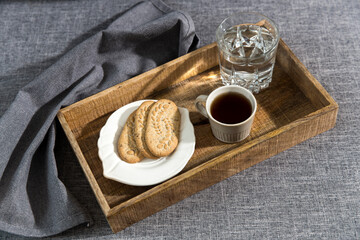 A white cup of coffee, a glass of water, a long cupronickel spoon with a twisted handle and a saucer with lemon for breakfast on a wooden tray, a rag napkin on a gray sofa.