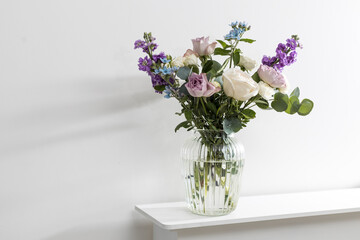 Bouquet of hackelia velutina, purple and white roses, small tea roses, matthiola incana and blue iris in glass vase is on the white coffee table.