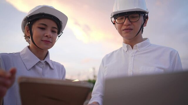 An architect foreman evaluating on a young female trainee's working knowledges, two engineer workers using electronic portable devices during late evening on outdoor workshop, coworker and team work