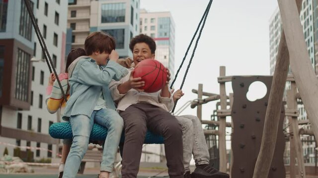 Slowmo shot of group of cheerful multiethnic school boys and girl having fun while swinging on nest swing seat outdoors at playground on sunny day