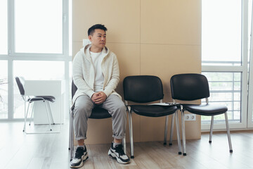 young asian man waiting for an interview or meeting sitting in the hallway in the waiting room. Student or entrant in the reception for exam or employment hr. Male patient in office a hospital clinic