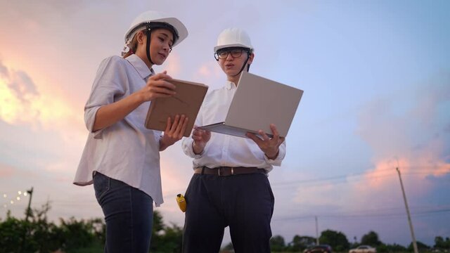 Asian Engineer architecture investor wear safety protective hard hat survey at outdoor construction site debriefing work during sunset, specialist expert working field, Real Estate industry innovation