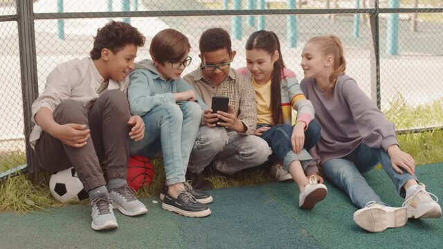 Slowmo shot of group of multiethnic school friends playing games on smartphone sitting on grass at playground on sunny day
