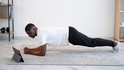Online training. Sportive black man. Strong body. Quarantine lifestyle. Happy african guy doing plank workout looking portable computer light room interior.