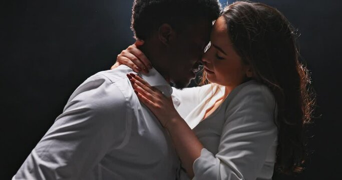 Close-up of dancing couple dressed in white, elegant woman holding partner by neck and with her other hand gently touching the man's chest, dark-skinned boy staring at his beloved, black background