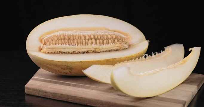 A cut melon is lying on a wooden cutting board. There are two melon slices nearby. Melon on a black background.