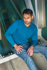 Dressing in a blue sweater and jeans , a young handsome guy with beard and mustache is sitting on the corner by a structure, bending over, looking down, lost in thought..