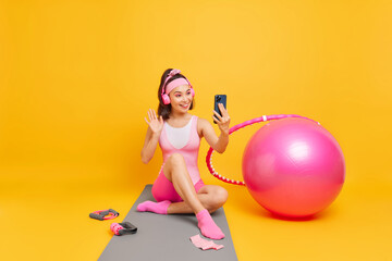 Fototapeta premium Positive smiling Asian woman with eastern appearance waves hello in smartphone camera has video call uses wireless headphones sits on karemat isolated over yellow background. Fitness training