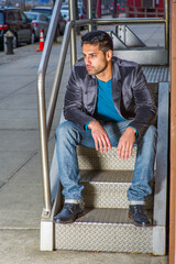 Dressing in a dark purple woolen blazer,  jeans and leather shoes , a young handsome worker with beard and mustache is sitting on door steps of an office building, bending over, relaxing.