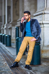 Fototapeta premium Dressing in blue woolen blazer, sweater, dark yellow pants and leather boot shoes, wearing glasses, a young guy with bread and mustache is sitting on an old fashion street, making a phone call.