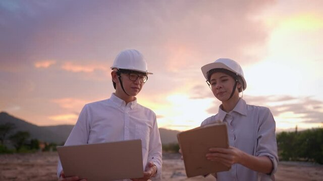 Asian Engineer and investor wear protective hardhat, using laptop and tablet talking on the project at outdoor site, colleague work environment, technology developer team, intern on the working site