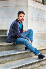 Dressing in a dark purple woolen blazer,  blue jeans and black leather shoes, a young handsome man is sitting on steps outside a office building, thinking