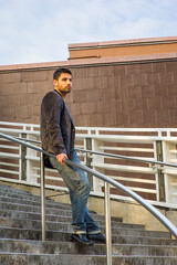 Dressing in dark purple woolen blazer, jeans and leather shoes, a young guy with beard and mustache is standing against a metal railing on stairs, under sunshine of sunset, thinking.