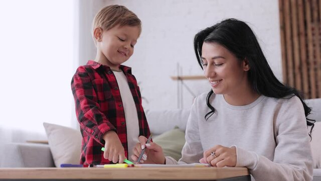 Smiling young caucasian mother teaching small cute child son drawing using colorful pencils in paper. Mom with child draw pictures with colored pencils. Enjoying spending free time together.
