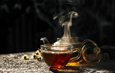 a cup of tea with steam with a glass teapot on a dark background 