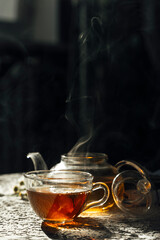 a cup of tea with steam with a glass teapot on a dark background 
