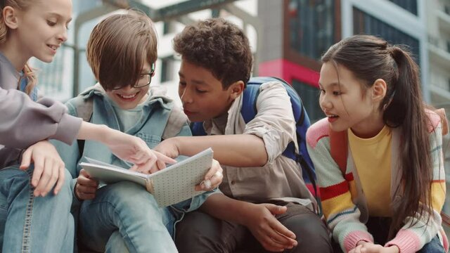 Slowmo shot of cheerful multiethnic group of school friends laughing while looking at copybook outdoors at schoolyard