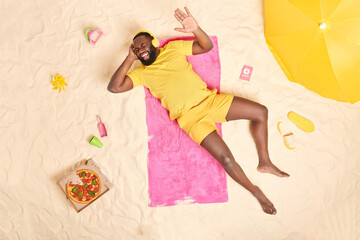 Positive dark skinned man in yellow clothing has fun listens music via headphones lies on pink soft towel spends free time at beach enjoys summer recreation eats tasty pizza drinks energetic beverage