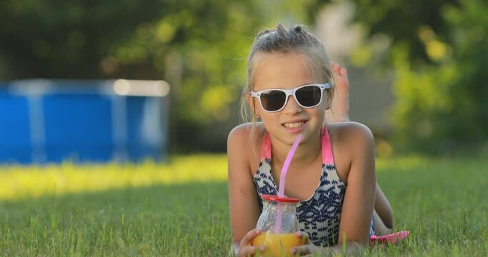 little girl in swimsuit and sunglasses drinks juice cocktail with drinking straw. Happy, cute child is lying on grass. Make faces and smile. Summer vacation concept