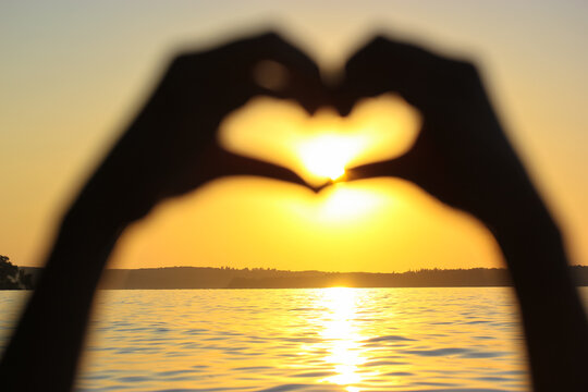 Silhouette of woman's hands in heart shape at sunset against a yellow-orange sky, sunbeams and a sun displayed inside. Symbol of love. Vacation by a sea, ocean, lake, river in summer. Seashore, coast.
