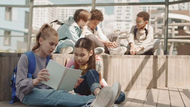 Slowmo shot of group of multiethnic school children resting outdoors in summer Girls discussing homework in copybook while boys playing games on smartphone