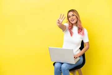 Young woman sitting on a chair with laptop over isolated yellow background happy and counting three with fingers