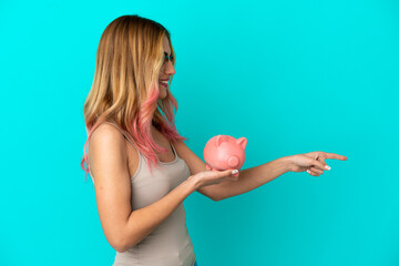 Young woman over isolated blue background holding a piggybank pointing to the side to present a product
