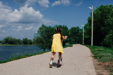 rear view of a girl in a yellow dress, a girl learns to ride a scooter in a natural park near the...