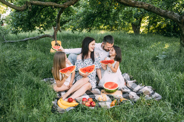 Young parents with two pretty daughters spending free time at green garden and eating tasty...