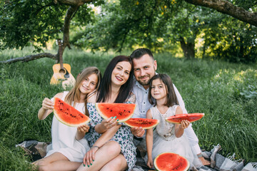 Caucasian couple with two little daughters sitting closely to each other at green garden with pieces of watermelon in hands. Positive family enjoying time spending together on picnic.