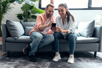 Happy young couple looking photos in smart phone while having fun sitting on couch at home.