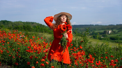 romantic girl in a red dress in a hat looks at the clear summer sky holding a poppy bouquet, wildflowers