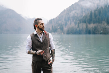 Fototapeta premium A brutal thoughtful young man groom with a dreadlocked hairstyle in a fashionable elegant wedding suit stands on the shore of a lake against the background of misty mountains in nature