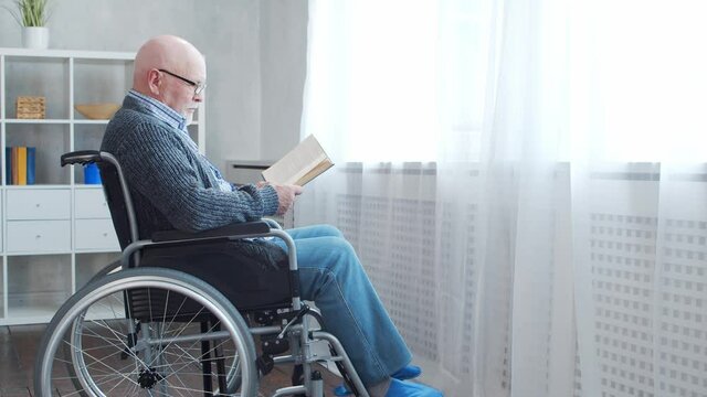 Disabled old man is sitting in a wheelchair at home alone. A handicapped person in a wheelchair is reading the book. Paralysis and disability.