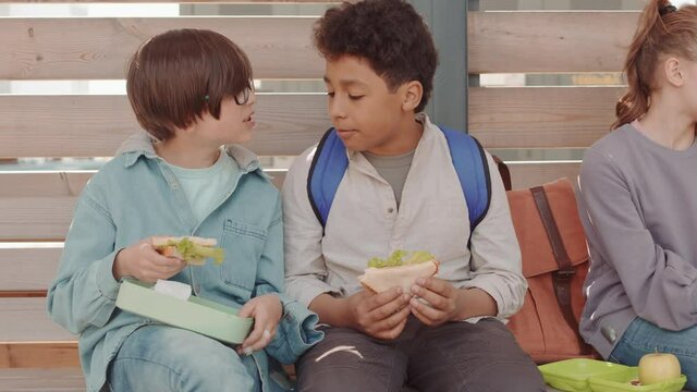 PAN medium slowmo shot of multiethnic group of school friends sitting on wooden bench at schoolyard eating sandwiches from lunchboxes between lessons