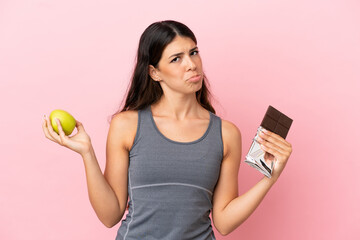 Young caucasian woman isolated on pink background having doubts while taking a chocolate tablet in...