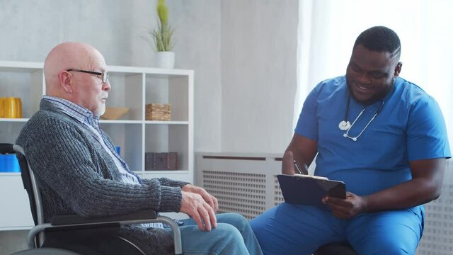 African-American caregiver and old disabled man in a wheelchair. Professional nurse and handicapped patient in a nursing home. Assistance, rehabilitation and health care.