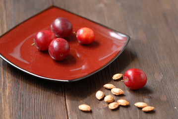 A red square dish with several fruits of red ripe cherry plums. Next to it on a wooden table there...