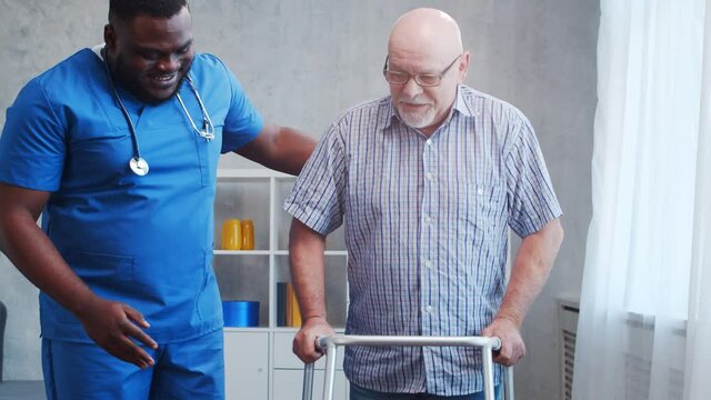 African-American caregiver is teaching disabled old man to walk with walker. Professional nurse and handicapped patient in a nursing home. Assistance, rehabilitation and health care.