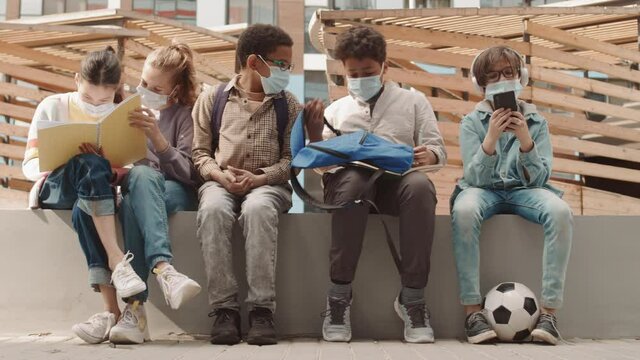 PAN slowmo shot of multiethnic school children in casual clothes and face masks resting after classes at playground or schoolyard sitting on bench together