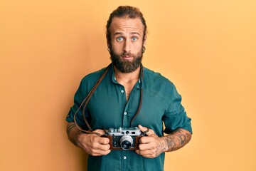 Handsome man with beard and long hair holding vintage camera clueless and confused expression....