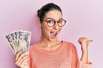 Young caucasian woman holding japanese yen banknotes pointing thumb up to the side smiling happy with open mouth