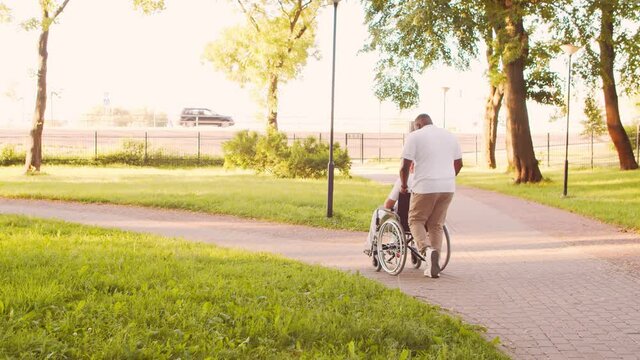 African-American caregiver and old disabled man in a wheelchair. Professional nurse and handicapped patient in the park. Assistance, rehabilitation and health care. Golden hour sunset.