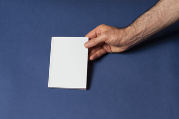 Male hand holding a closed notebook with blank cover on blue background - editable mockup template