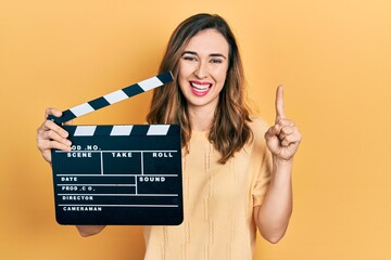 Young hispanic girl holding video film clapboard smiling with an idea or question pointing finger...