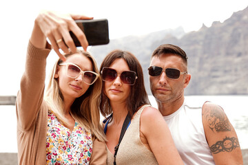 Three smiling friends taking selfie photos at digital camera of smartphone. Summer time.